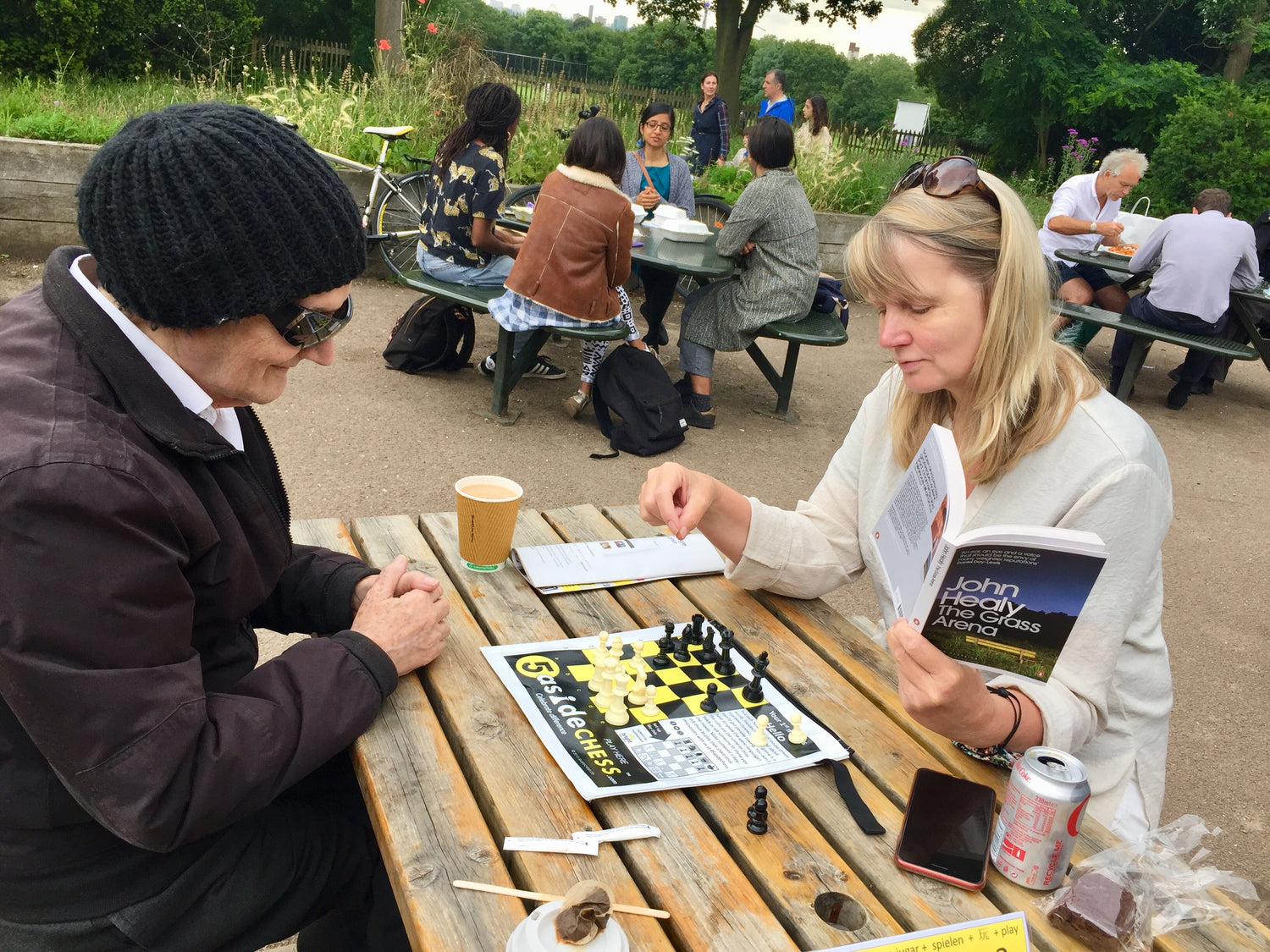 Two people playing 5asideCHESS at a picnic table in an outdoor setting with other people in the background.
