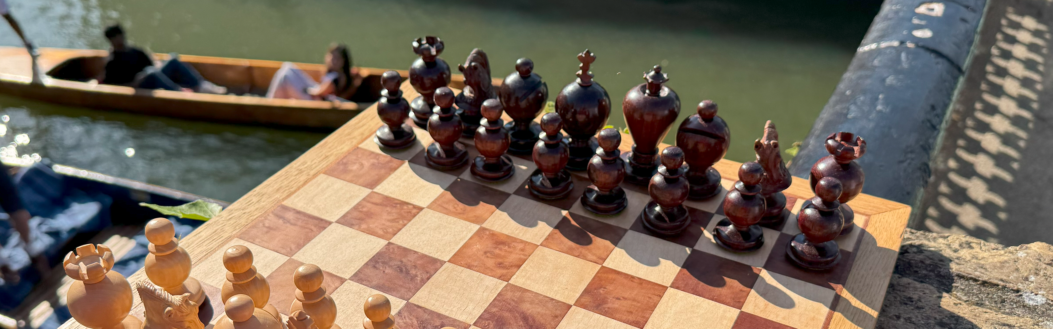 a vintage chess board and pieces with a background of the river Cam and people in a punt.