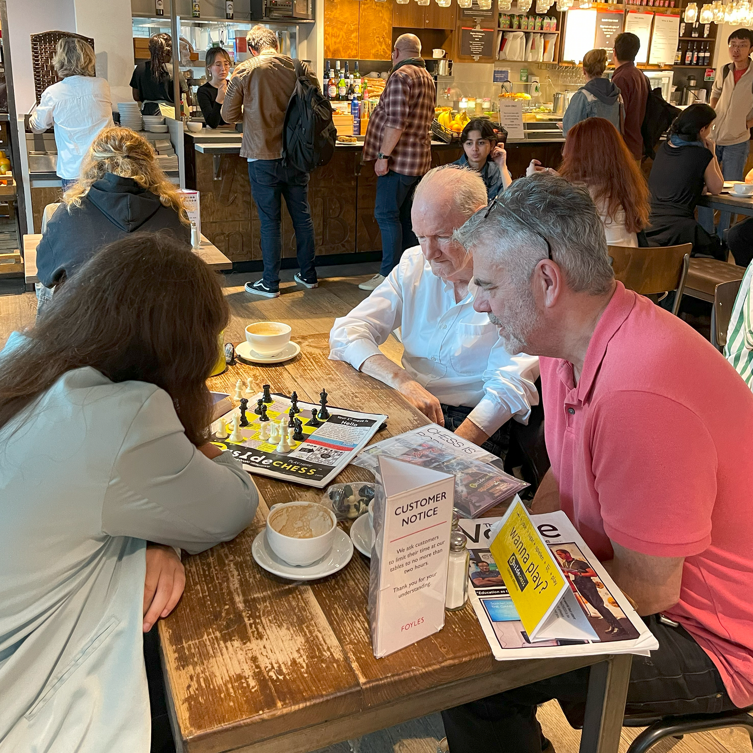 Two men playing 5asideCHESS at a table in a busy cafe with other patrons in the background.